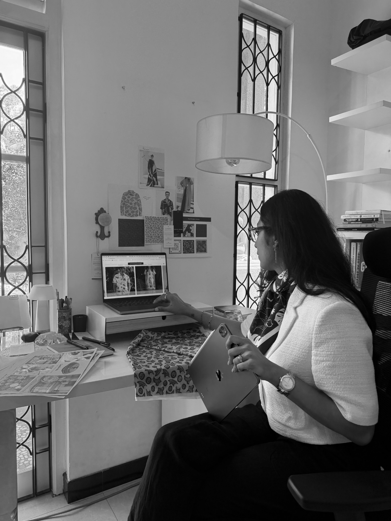 Black-and-white portrait of Celeste at her studio desk with a tablet, looking at the collection on her laptop beneath the moodboard