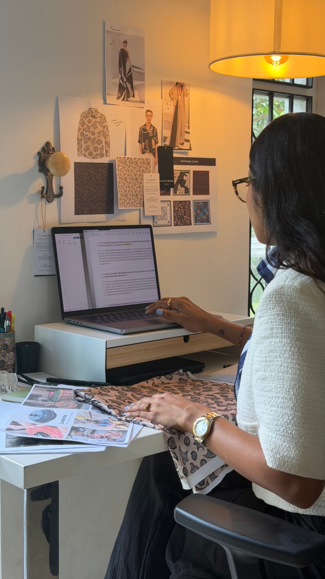 Celeste at her studio desk under a warm lamp, working on the LAAKSHA collection with print designs pinned on the wall and fabric across the desk